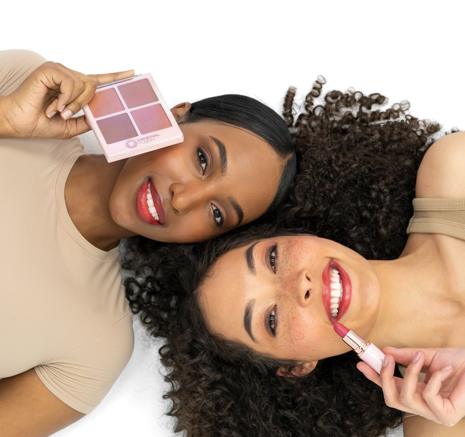 Two women lying down, one holding a makeup palette and the other with a lip product, on a white background.